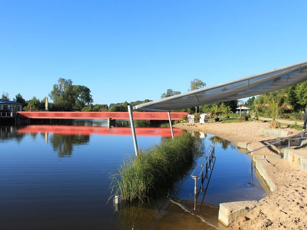 Kneippbecken Auf dem Bild ist ein See mit roter Brücke, Sandstrand und grüner Umgebung unter klarem Himmel.
