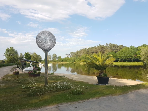 Ruhiger See mit gepflegtem Uferweg, Skulptur und Palmentopf, umgeben von Bäumen unter blauem Himmel.
