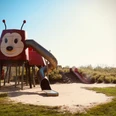 Roter Spielplatz in Form einer Ameise mit Rutsche und Sandfläche, umgeben von grüner Vegetation.