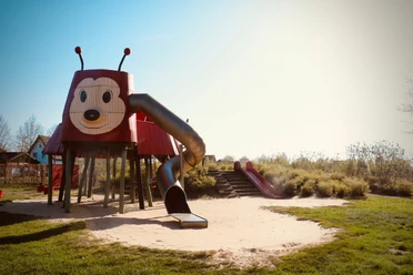 Rieti-Rutsche Roter Spielplatz in Form einer Ameise mit Rutsche und Sandfläche, umgeben von grüner Vegetation.