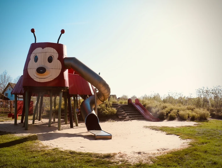 Rieti-Rutsche Roter Spielplatz in Form einer Ameise mit Rutsche und Sandfläche, umgeben von grüner Vegetation.
