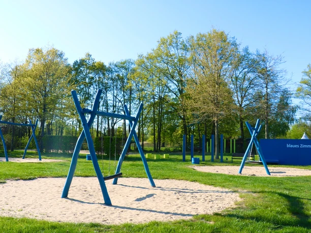 Ein Spielplatz im Freien mit mehreren blauen Schaukeln und Klettergeräten auf Sandflächen.