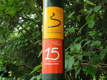 Markierungszeichen Bergbauweg Wegweiser eines Wanderwegs mit gelbem und rotem Schild, umgeben von dichtem Laubwald.