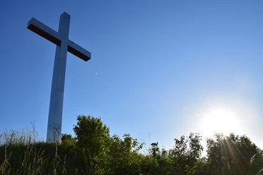 Barbarakreuz <p>Großes weißes Kreuz auf einem Hügel, umgeben von Büschen, mit strahlend blauem Himmel und Sonne.</p>