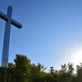 Barbarakreuz <p>Großes weißes Kreuz auf einem Hügel, umgeben von Büschen, mit strahlend blauem Himmel und Sonne.</p>