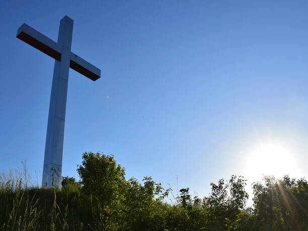 Barbarakreuz <p>Großes weißes Kreuz auf einem Hügel, umgeben von Büschen, mit strahlend blauem Himmel und Sonne.</p>