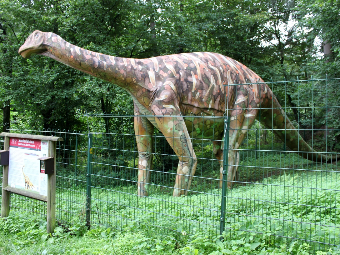 Modell eines Megalosauriers an den Saurierfährten in Bad Essen – Barkhausen. Dinosauriermodell in einem grünen Park hinter einem Metallzaun, neben einem Infotafel.Dinosaur model in a green park behind a metal fence, next to an information board.Dinosaurmodel i en grøn park bag et metalhegn ved siden af en informationstavle.Dinosaurusmodel in een groen park achter een metalen hek, naast een informatiebord.