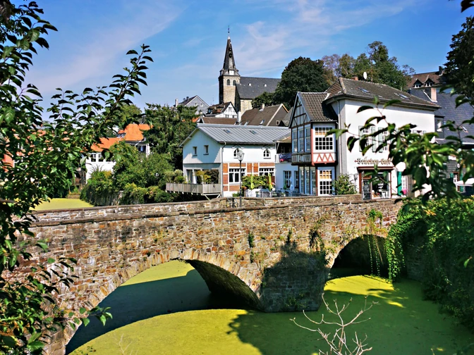 RevierRoute GartenStadt.jpg <p>Historische Steinbrücke über grün schimmerndem Wasser, dahinter Fachwerkhäuser und ein Kirchturm.</p>