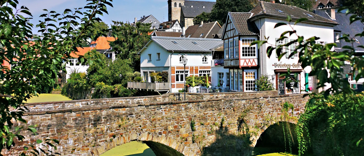 RevierRoute GartenStadt <p>Historische Steinbrücke über grün schimmerndem Wasser, dahinter Fachwerkhäuser und ein Kirchturm.</p>