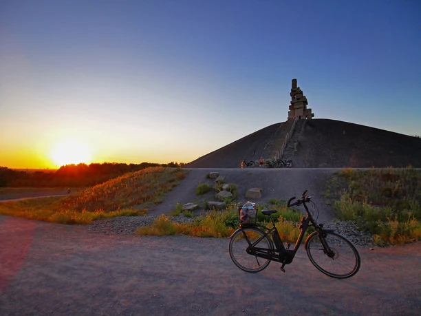 Ein einsames Fahrrad steht bei Sonnenuntergang vor einer Haldenlandschaft mit einem markanten Gipfelbau.