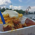 RevierRoute Zweistromland.JPG Pommes mit Mayonnaise und Currywurst in einer Plastikschale vor einem See mit Kran und Bewölkung.