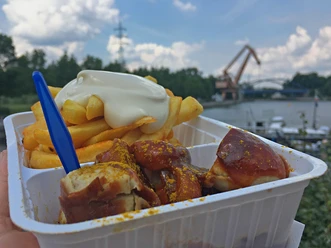 RevierRoute Zweistromland Pommes mit Mayonnaise und Currywurst in einer Plastikschale vor einem See mit Kran und Bewölkung.