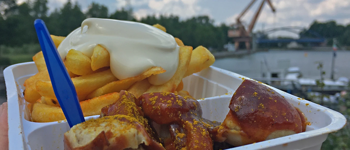 RevierRoute Zweistromland Pommes mit Mayonnaise und Currywurst in einer Plastikschale vor einem See mit Kran und Bewölkung.
