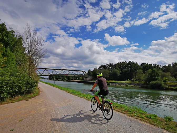 RevierRoute Kanalpassage.jpg Radfahrer auf einem sonnigen Pfad am Fluss neben einer grünen Landschaft und Brücke.