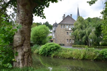 Ensemble Volberg Eine historische Steinmühle an einem kleinen Fluss, umgeben von üppiger Vegetation und Bäumen.