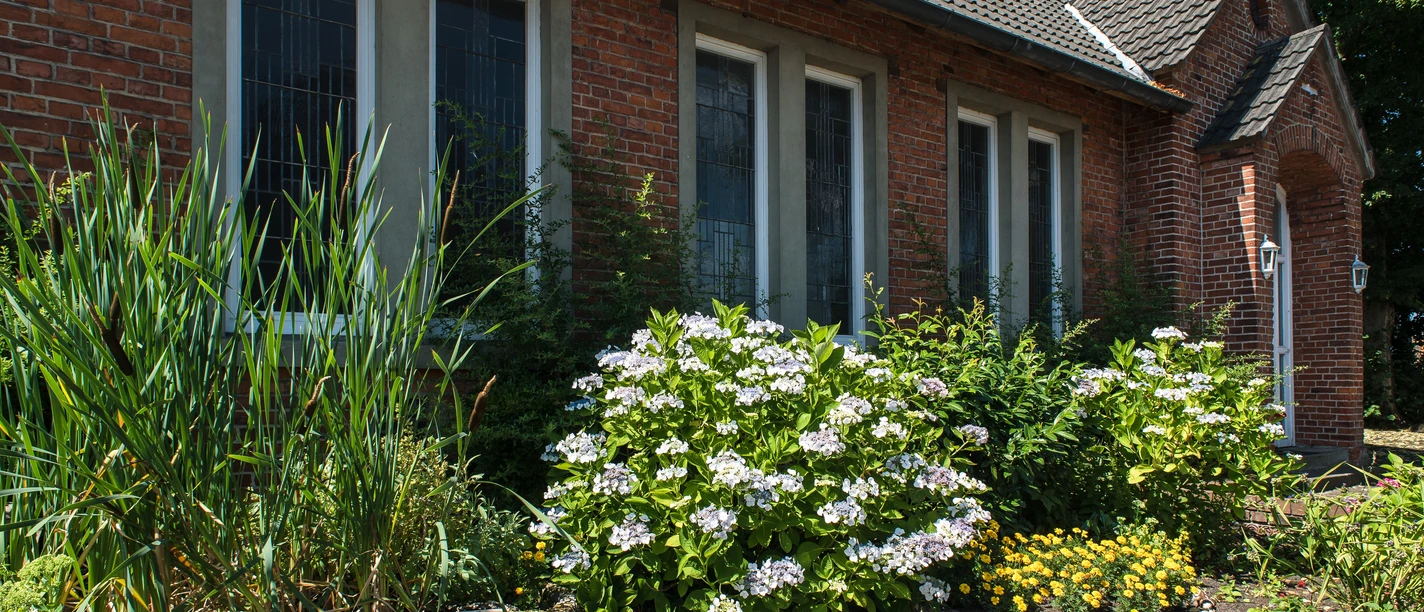 Alte Schule Spelle Backsteingebäude mit hohen Fenstern und gepflegtem Vorgarten voller blühender Sträucher und Pflanzen.