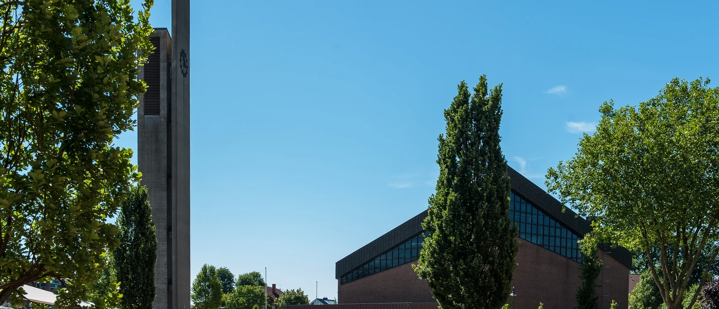 Kath. Kirsche St. Johannes der Täufer Moderner Kirchenbau mit hohem Glockenturm, Brunnen und Figurengruppe auf einem sonnigen Platz.