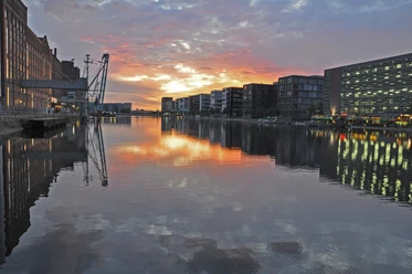 Duisburg Innenhafen Abendstimmung.JPG <p>Sonnenuntergang spiegelt sich im ruhigen Wasser eines Hafenbeckens, umgeben von Gebäuden und Kränen.</p>