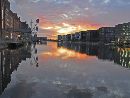 Duisburg Innenhafen Abendstimmung.JPG <p>Sonnenuntergang spiegelt sich im ruhigen Wasser eines Hafenbeckens, umgeben von Gebäuden und Kränen.</p>