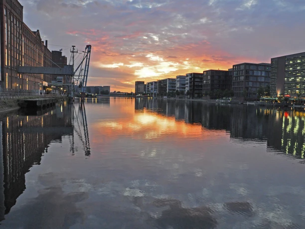 Duisburg Innenhafen Abendstimmung.JPG <p>Sonnenuntergang spiegelt sich im ruhigen Wasser eines Hafenbeckens, umgeben von Gebäuden und Kränen.</p>