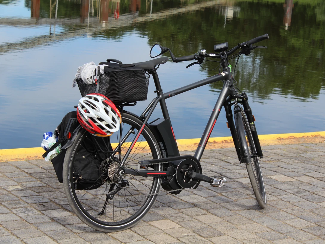 Ein schwarzes Fahrrad mit Gepäckträger und Helm steht auf gepflastertem Boden vor ruhigem Wasser.
