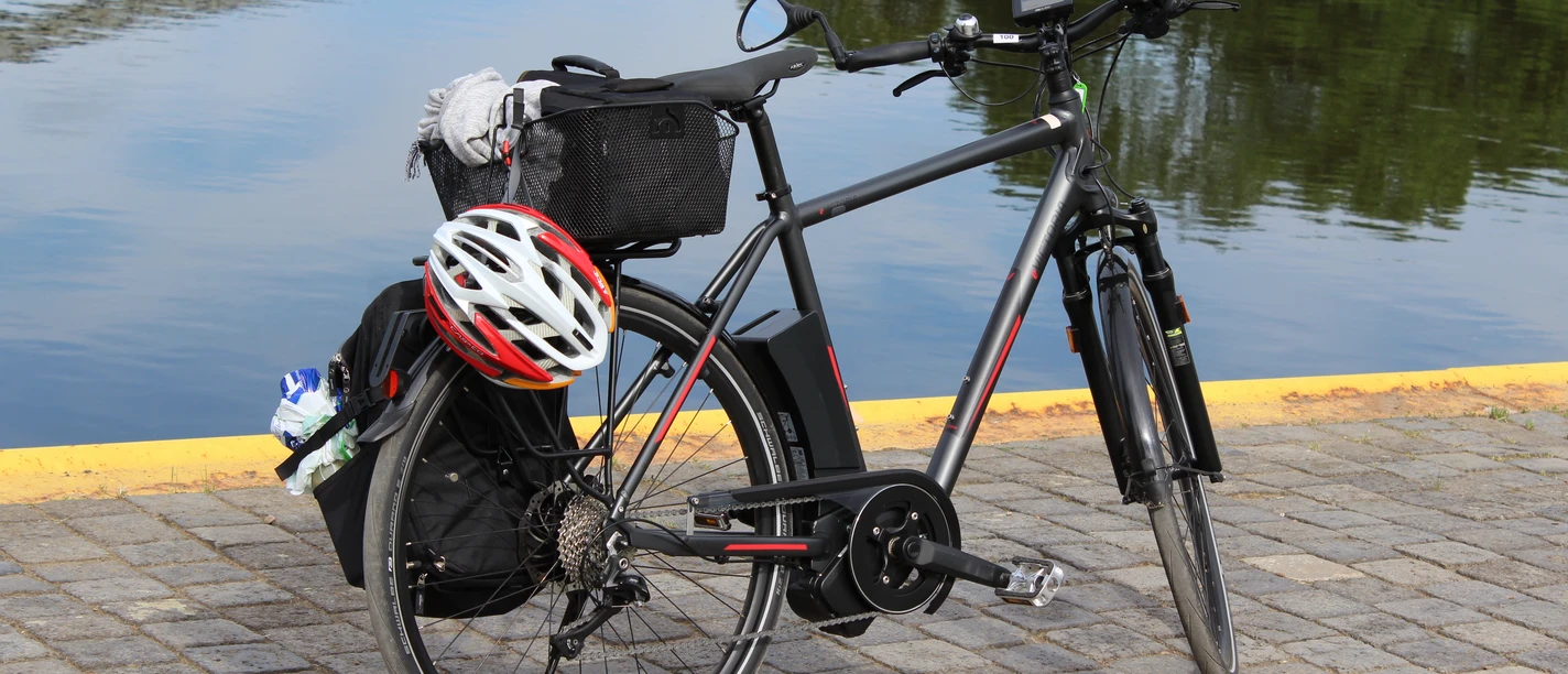 Ein schwarzes Fahrrad mit Gepäckträger und Helm steht auf gepflastertem Boden vor ruhigem Wasser.