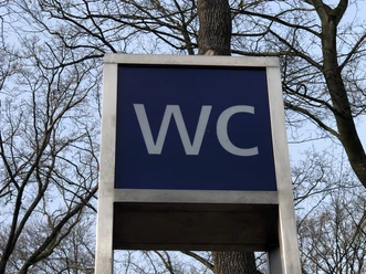 Öffentliche Toilette in einem parkähnlichen Gebiet, umgeben von Bäumen, unter freiem Himmel.