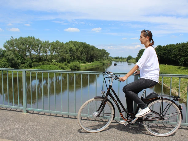A woman on a bicycle enjoys the view of the Weser from a bridge. The surroundings are green and peaceful.