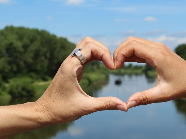 Two hands form a heart in front of a river landscape, with trees and blue sky in the background.