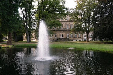 Palaisgarten Brunnen mit sprudelndem Wasser vor einem historischen Gebäude, umgeben von grünen, großen Bäumen.