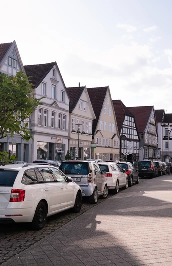 Straßenansicht mit parkenden Autos und traditionellen Fachwerkhäusern unter wolkigem Himmel.