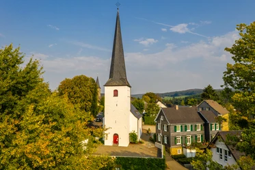 ev. Kirche Ruppichteroth Altstadt <p>Weiße Kirche mit hohem Turm zwischen Bäumen, flankiert von traditionellen Fachwerkhäusern.</p>