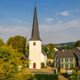 ev. Kirche Ruppichteroth Altstadt <p>Weiße Kirche mit hohem Turm zwischen Bäumen, flankiert von traditionellen Fachwerkhäusern.</p>