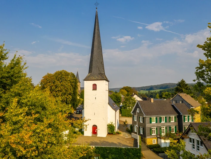 ev. Kirche Ruppichteroth Altstadt <p>Weiße Kirche mit hohem Turm zwischen Bäumen, flankiert von traditionellen Fachwerkhäusern.</p>