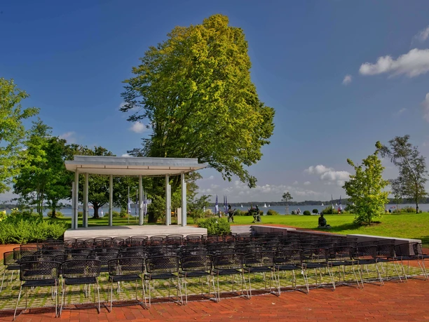 freilichtbühne-kurpark-mit-stuehlen.jpg Die Freilichtbühne im Kurpark vor zahlreichen leeren Metallstühlen, umgeben von grünem Rasen und Bäumen, mit einem malerischen Blick auf einen See im Hintergrund und einigen Spaziergängern in der Ferne.