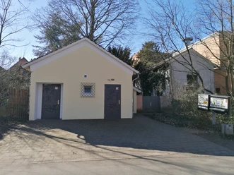 Öffentliche Toiletten mit zwei separaten Eingängen, umgeben von Bäumen, auf gepflastertem Boden.
