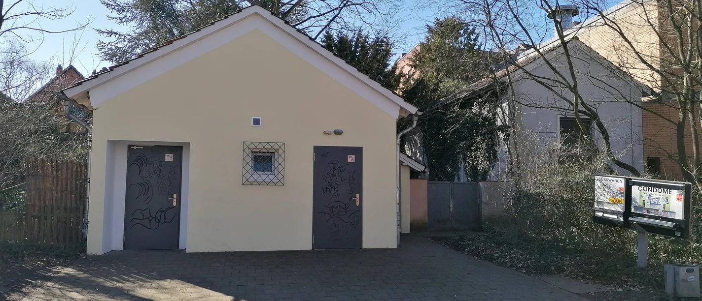 Öffentliche Toiletten mit zwei separaten Eingängen, umgeben von Bäumen, auf gepflastertem Boden.