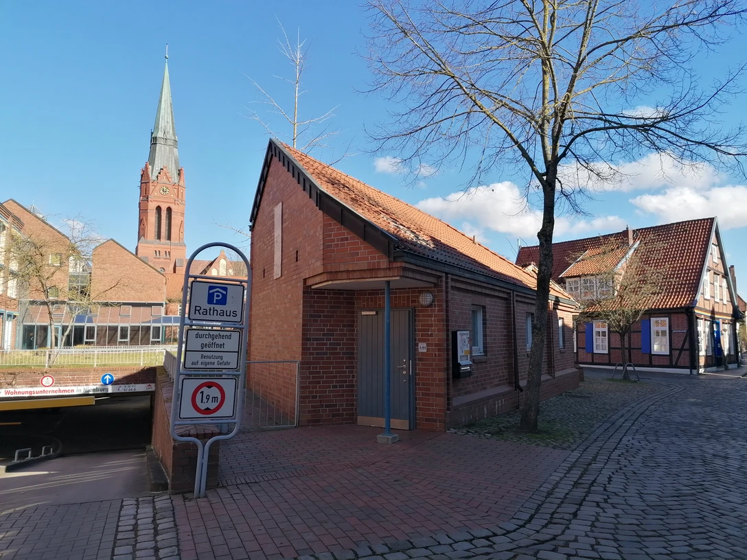 Rote Backstein-Toilettenanlage in der Nähe des Rathauses, mit Zugang zur Tiefgarage und Kirchturm im Hintergrund.
