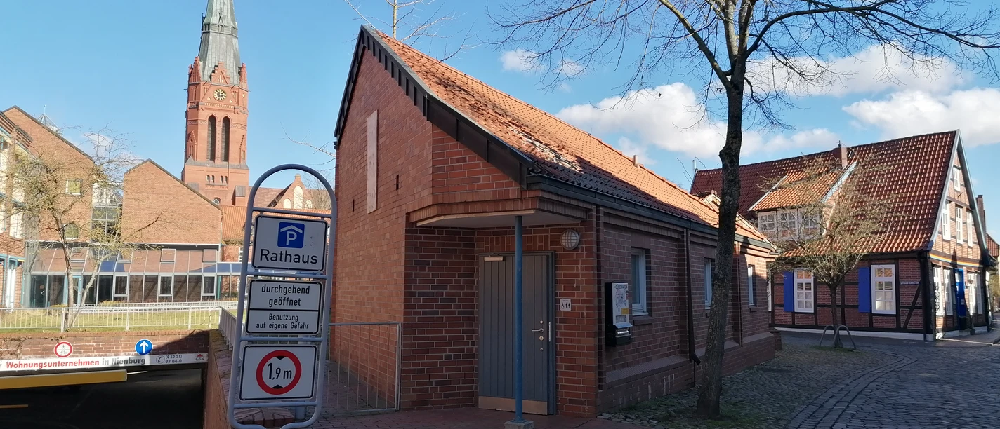 Rote Backstein-Toilettenanlage in der Nähe des Rathauses, mit Zugang zur Tiefgarage und Kirchturm im Hintergrund.