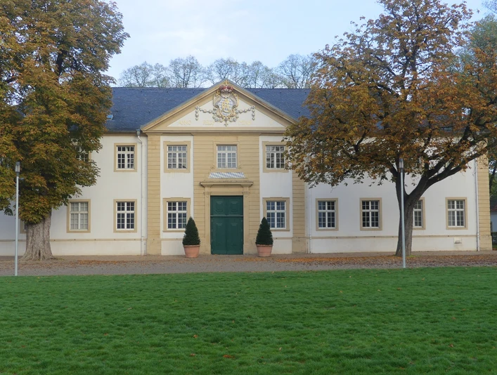Ehemaliger Marstall mit weißer Fassade und grünem Tor, flankiert von Herbstbäumen auf Rasenfläche.