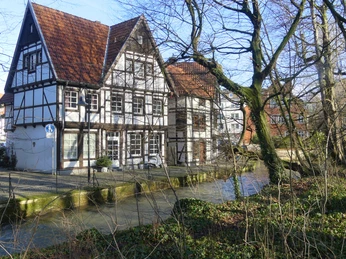 Fachwerhäuser Auf den Dielen Historische Fachwerkhäuser mit roten Ziegeldächern, umgeben von fließendem Bach und winterlichen Bäumen.