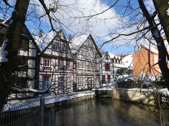 Fachwerhäuser Auf den Dielen - Winterbild Verschneite Fachwerkhäuser säumen einen ruhigen Bachlauf, umgeben von kahlen Bäumen an einem klaren Wintertag.