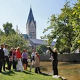 Eine Gästeführerin erklärt einer Gruppe die Paderquellen in Paderborn, im Hintergrund ein Kirchturm.