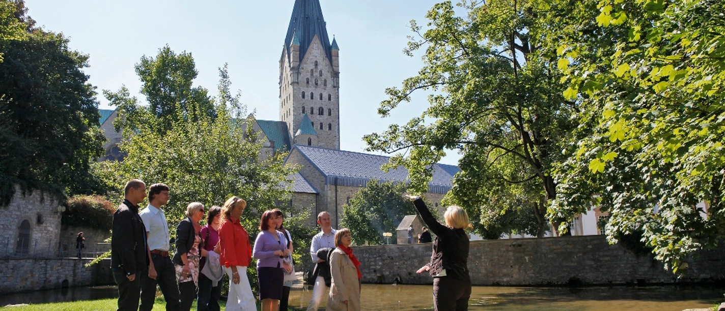 Eine Gästeführerin erklärt einer Gruppe die Paderquellen in Paderborn, im Hintergrund ein Kirchturm.
