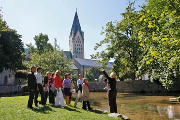 Paderquellen Paderborn (Gästeführung) Eine Gästeführerin erklärt einer Gruppe die Paderquellen in Paderborn, im Hintergrund ein Kirchturm.