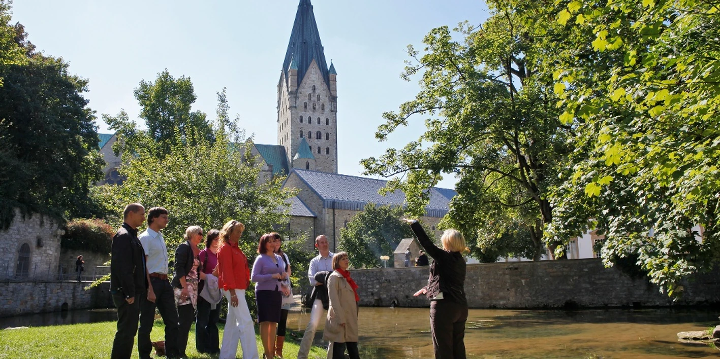 Paderquellen Paderborn (Gästeführung) Eine Gästeführerin erklärt einer Gruppe die Paderquellen in Paderborn, im Hintergrund ein Kirchturm.