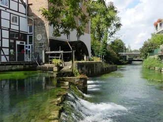 Historische Mühle am klaren Fluss mit hölzernem Wasserrad, umgeben von Fachwerk und Natur.