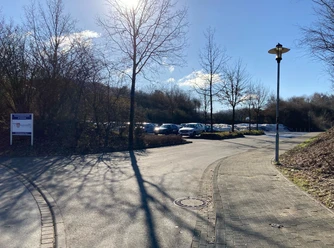 Gepflasterter Weg mit Straßenlaterne führt zu einem Parkplatz, Bäume und blauer Himmel im Hintergrund.