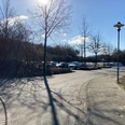 Gepflasterter Weg mit Straßenlaterne führt zu einem Parkplatz, Bäume und blauer Himmel im Hintergrund.