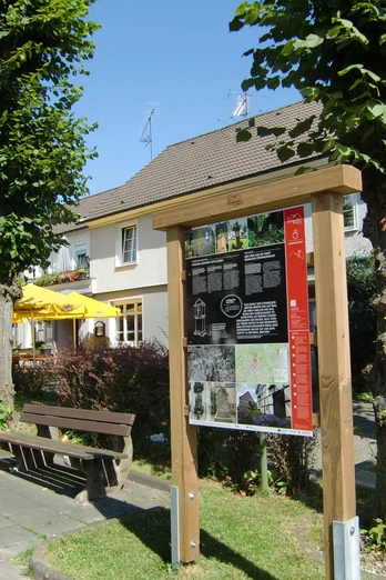 Infotafel am Steinhauerpfad Ein Infobrett neben Parkbänken und Bäumen, im Hintergrund ein Gebäude mit gelben Sonnenschirmen.